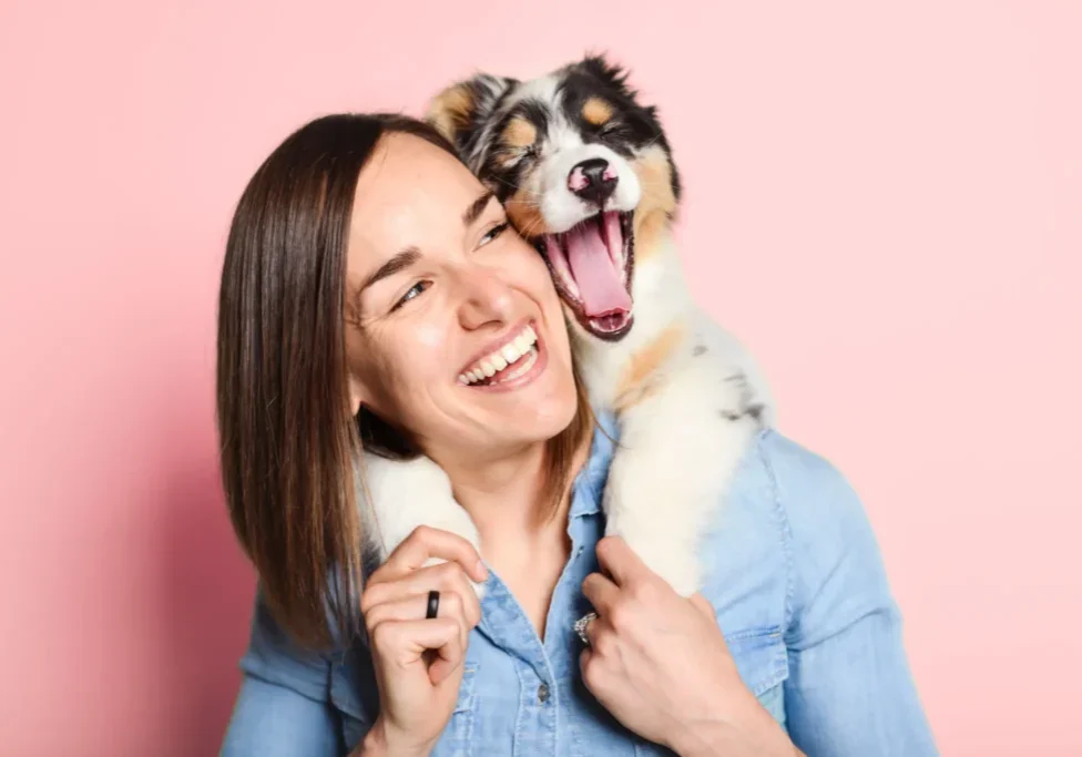 A joyful woman holding a happy puppy against a pink background.